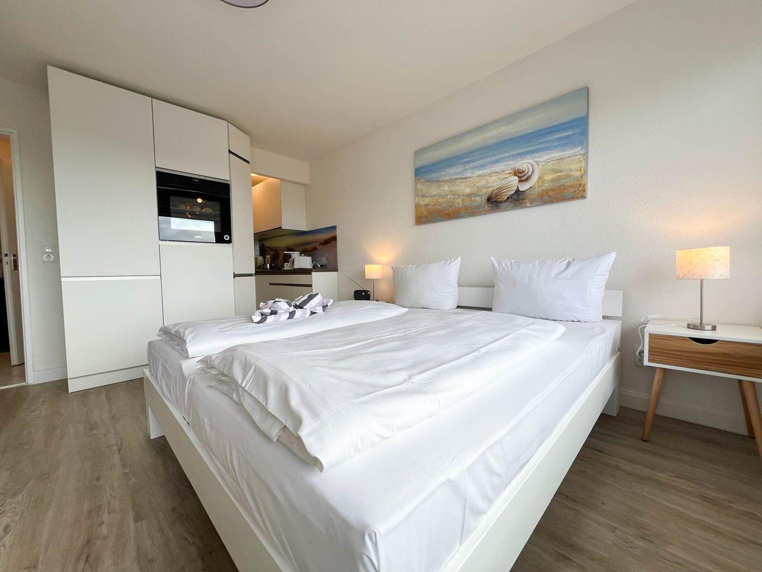 Schlafzimmer mit Bett, Nachttisch und Küchenbereich im Hintergrund.