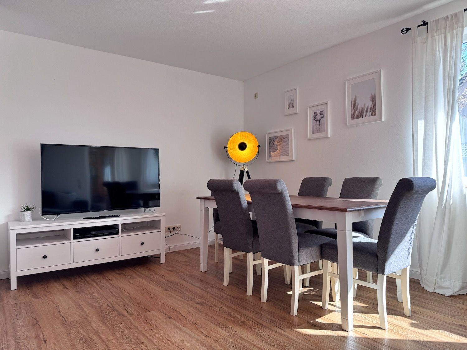 Wohn- und Essbereich mit TV, Esstisch und Stühlen. Holzboden und Wandbilder.