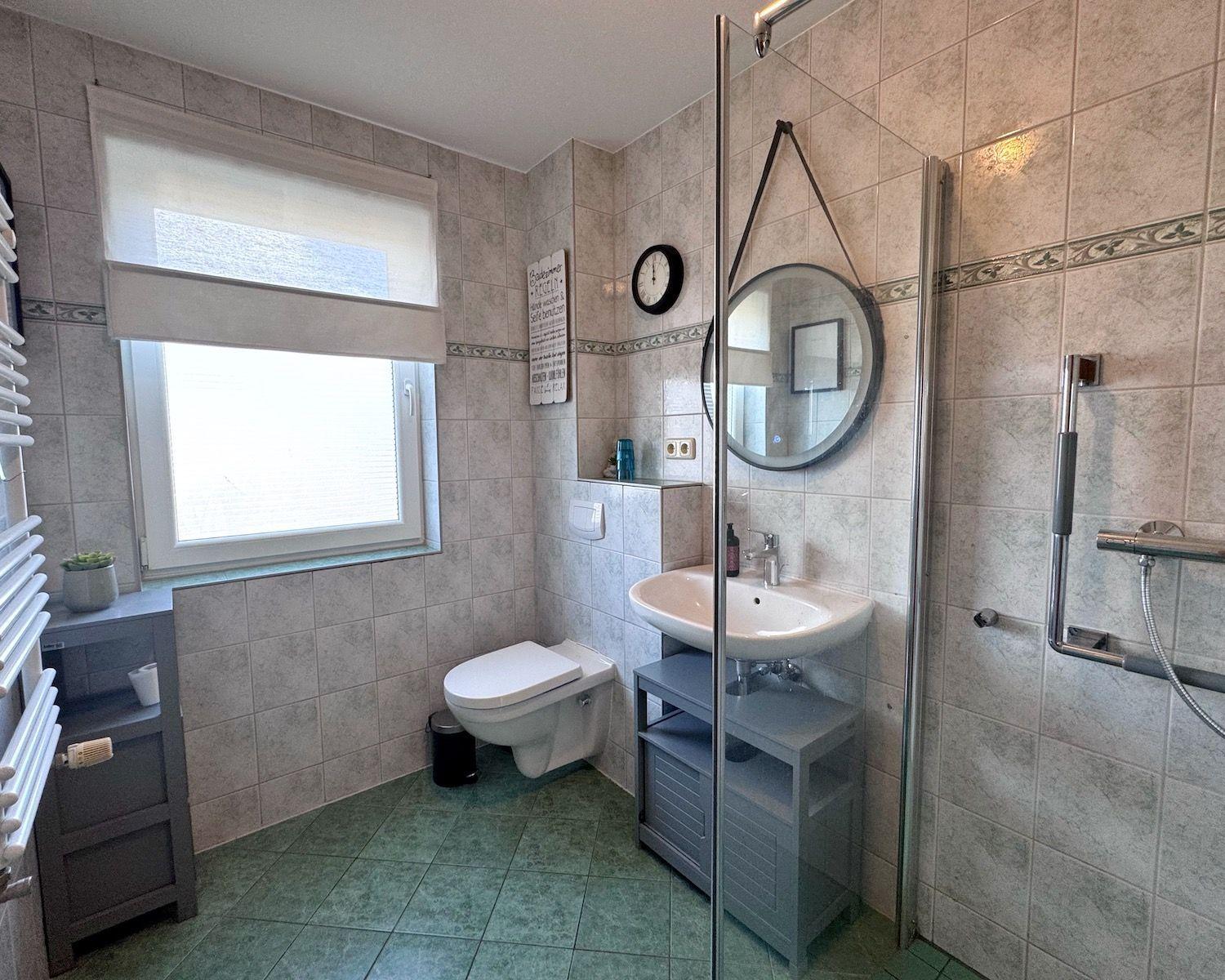 Badezimmer mit Dusche, Waschbecken und Toilette. Fenster mit Rollladen und Wanduhr.
