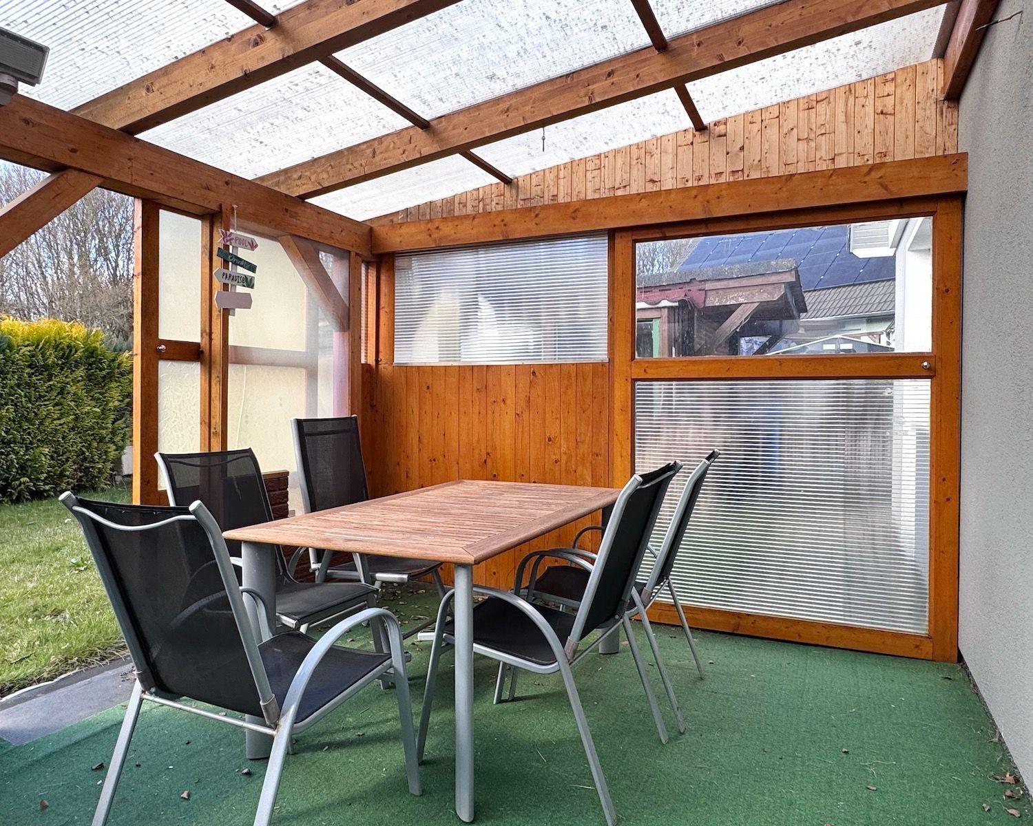 Terrasse mit Tisch und Stühlen unter Holzkonstruktion mit transparentem Dach.