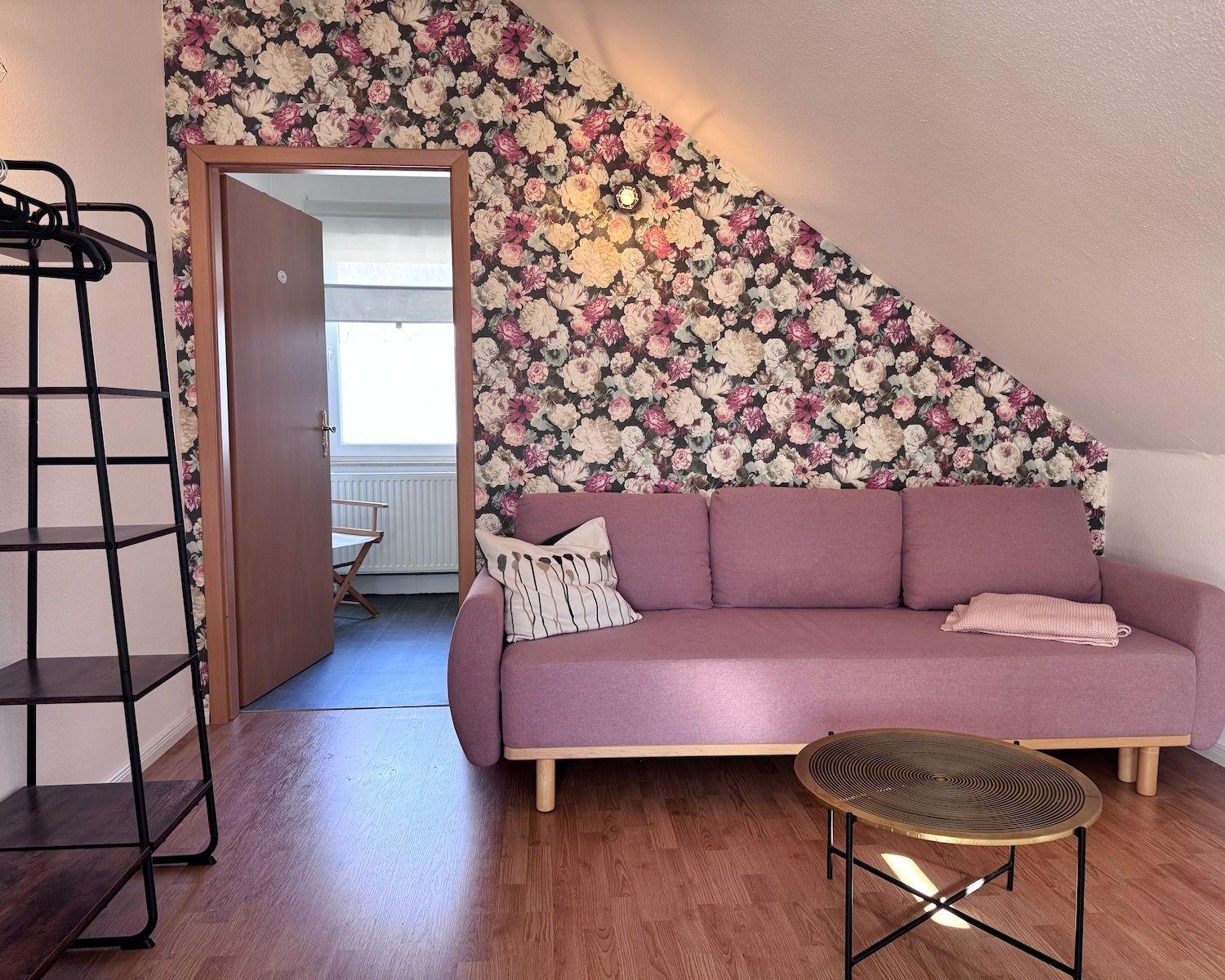 Ein Raum mit rosa Sofa, floraler Wandtapete und Holzboden. Eine Tür führt in einen anderen Raum.