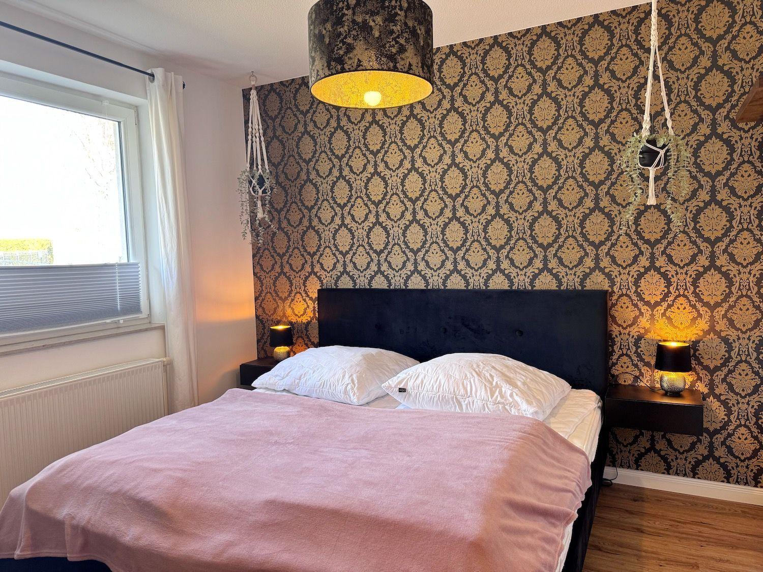 Schlafzimmer mit Bett, rosa Bettwäsche, dunkler Tapete und hängenden Pflanzen.