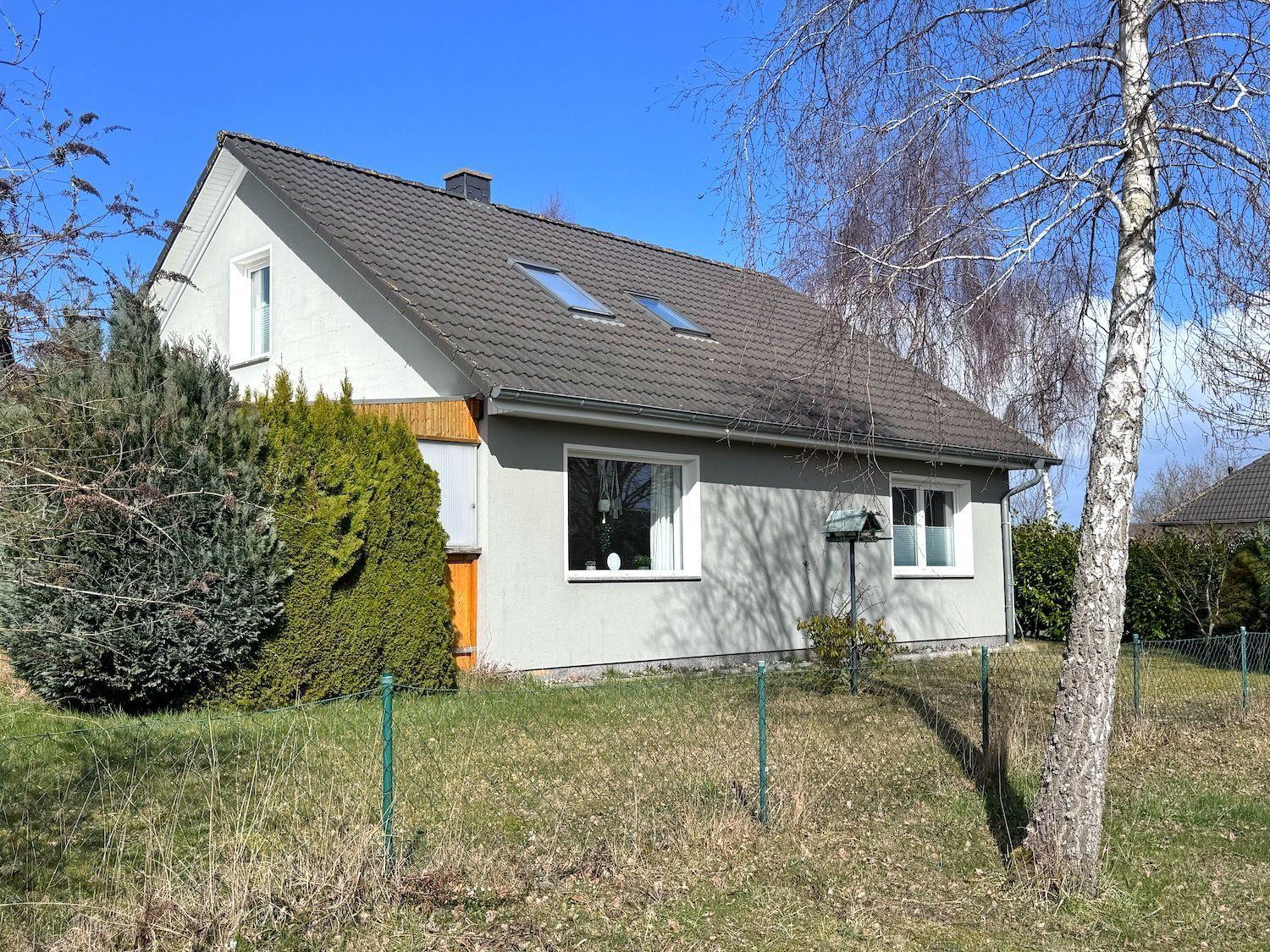 Ein zweistöckiges Haus mit grauer Fassade, Dachgauben und Garten mit Bäumen.