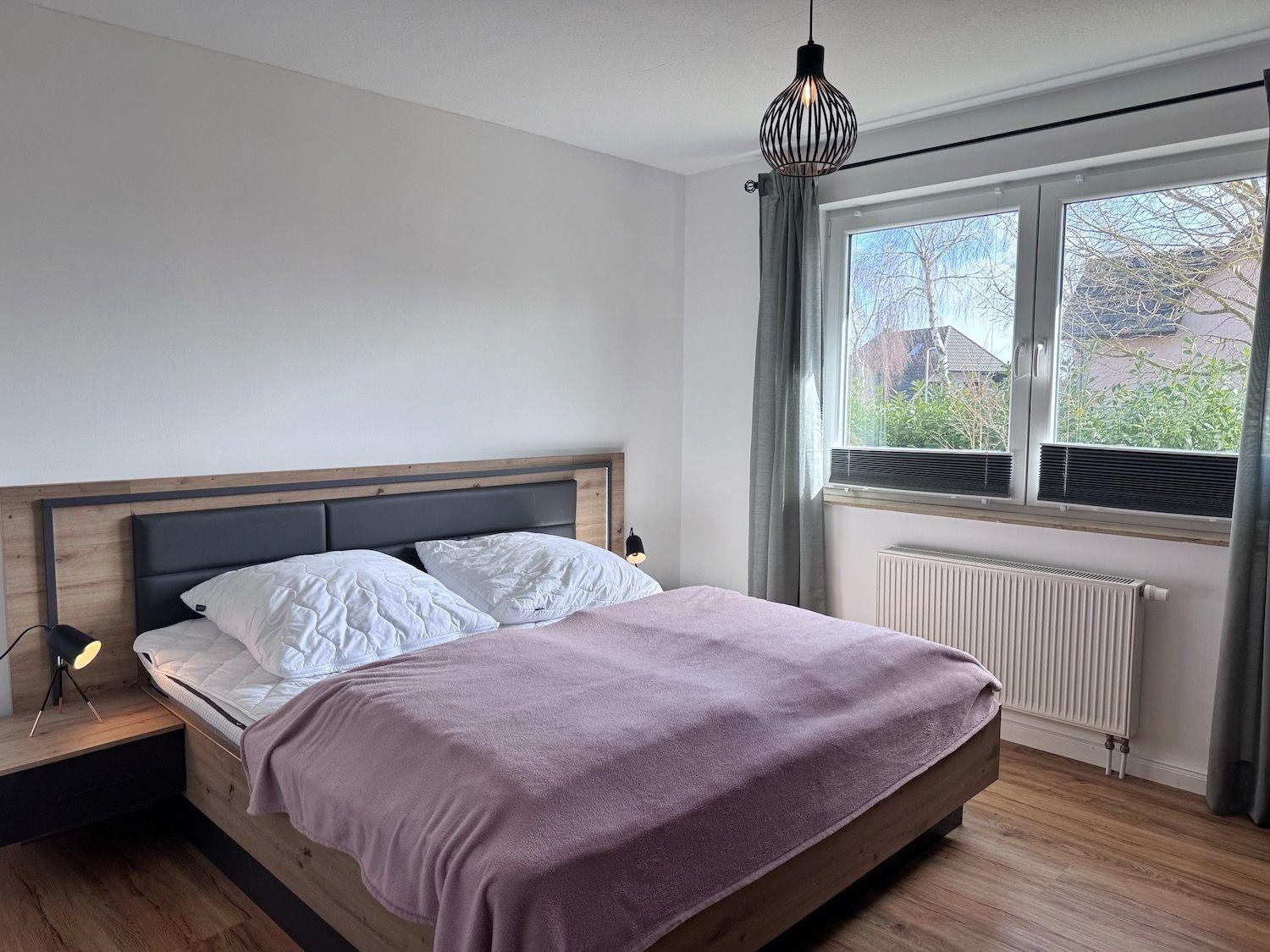 Schlafzimmer mit Doppelbett, Holzboden und Fenster mit Blick auf Außenbereich.