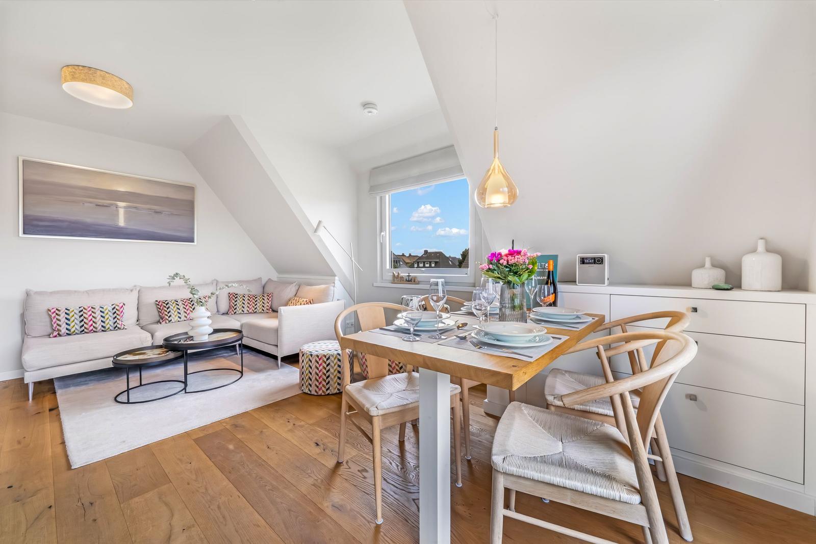 Living and dining area with sofa, table, and chairs. Wooden floor and large windows.