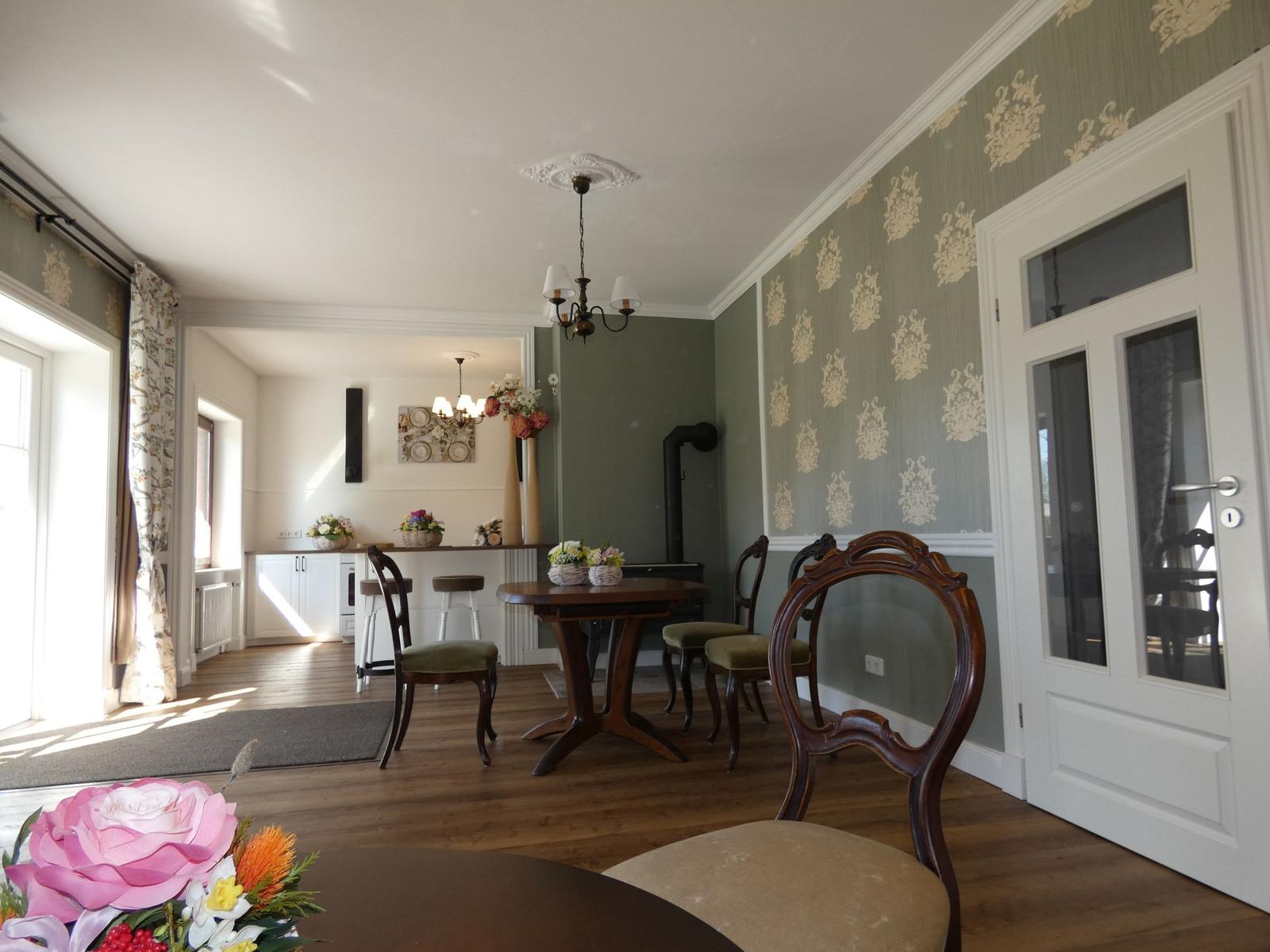 Esszimmer mit Holztisch, Stühlen und floraler Tapete. Küche im Hintergrund.