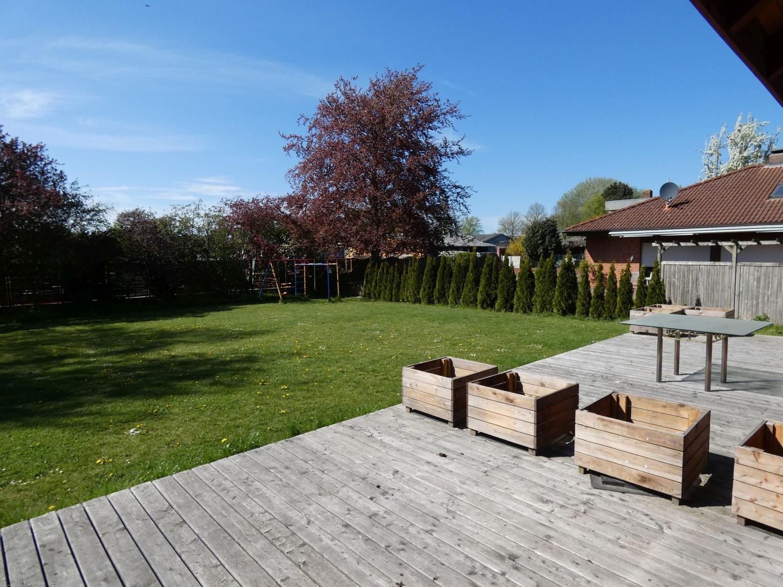 Holzterrasse mit Tisch und Kisten, grüner Garten mit Spielplatz und Bäumen.