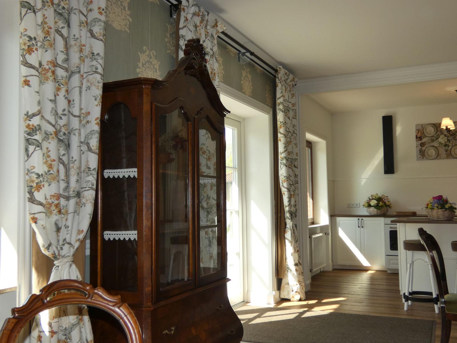 Ein Raum mit einem antiken Schrank, floralen Vorhängen und Blick in die Küche.