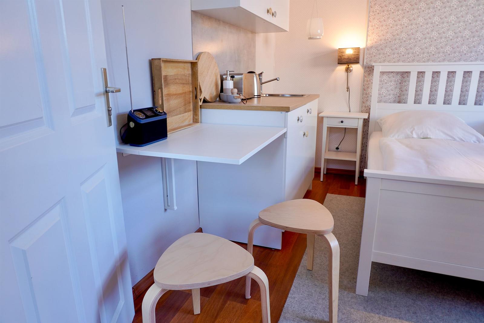 Kleine Küche mit Arbeitsfläche, Bett und Nachttisch. Holzstühle und Wandlampe sichtbar.
