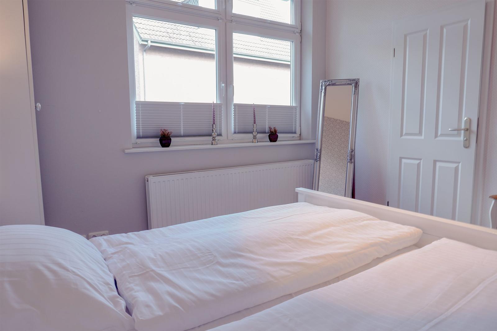 Schlafzimmer mit weißem Bett, Fenster mit Jalousien und Spiegel an der Wand.