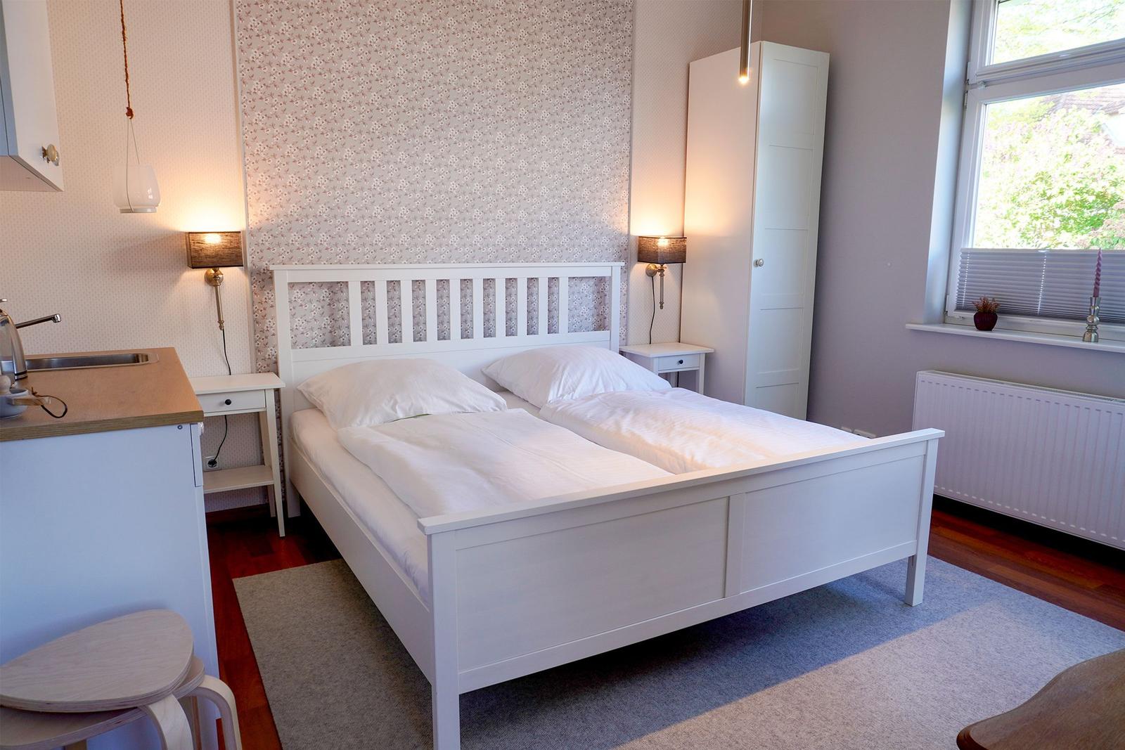 Schlafzimmer mit weißem Bett, Nachttischen und Fenster. Hinter dem Bett ist ein gemustertes Wandpaneel.