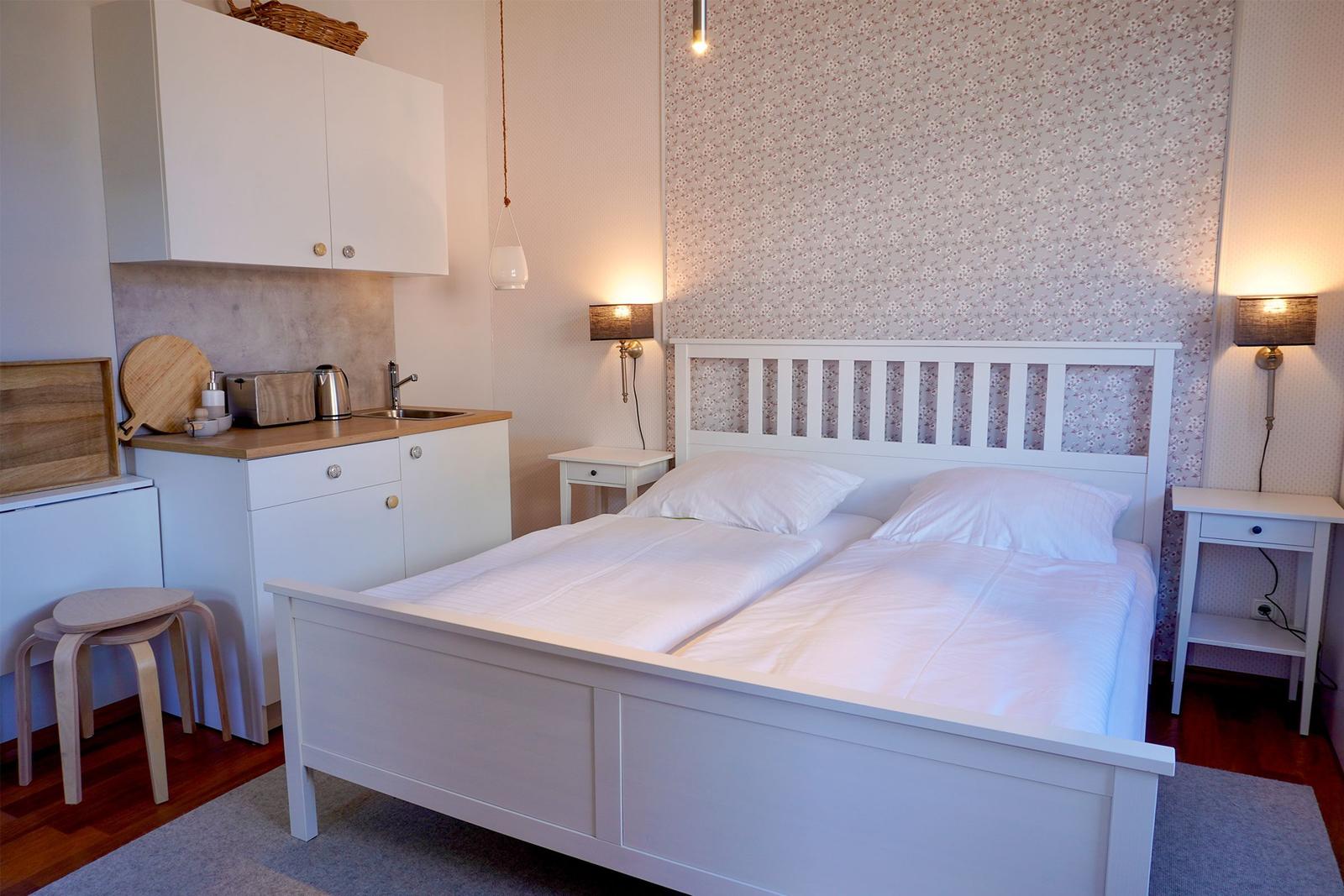 Ein Schlafzimmer mit weißem Bett, Nachttischen und kleiner Küchenzeile.