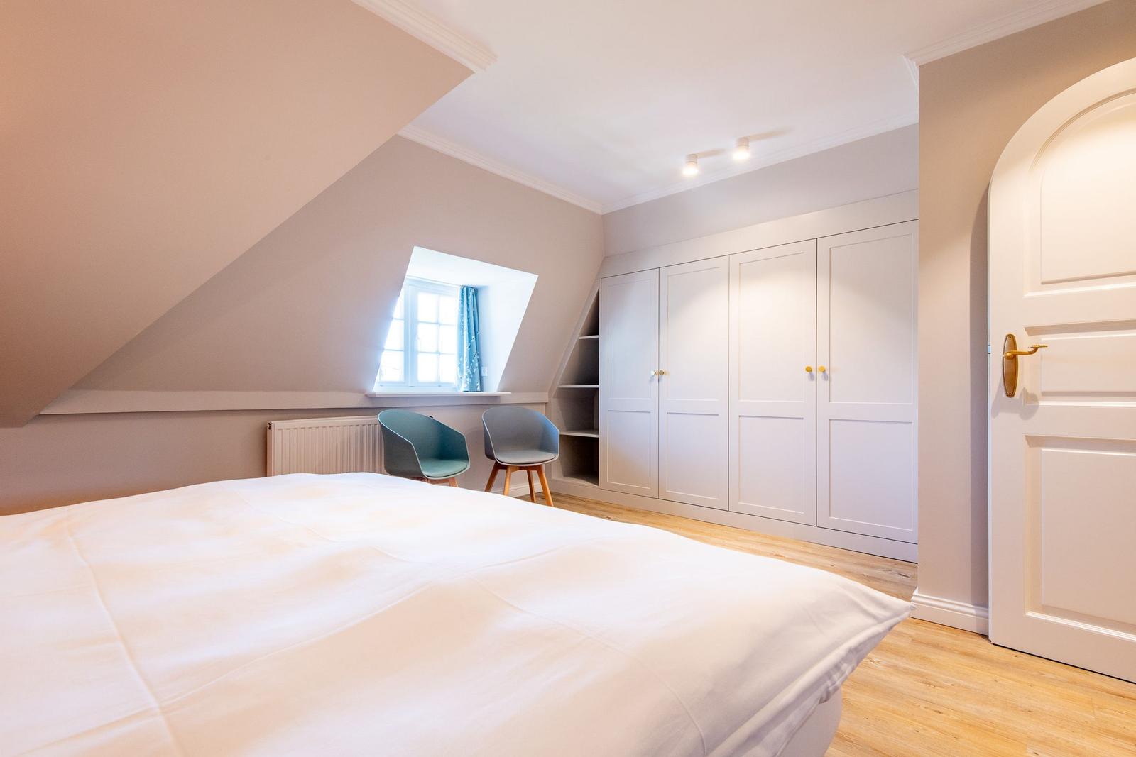 Bedroom with sloped ceilings, white duvet, two chairs, and white wardrobes.