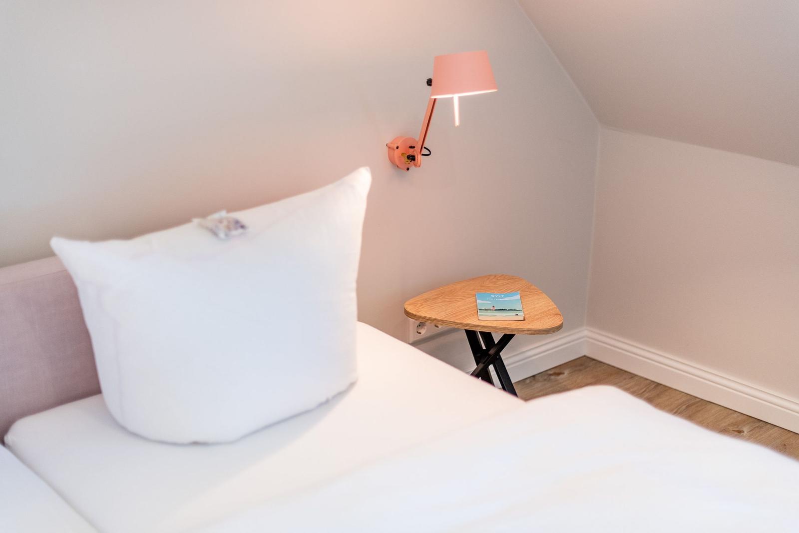 A bed with white linens, a pink wall lamp, and a small wooden table.