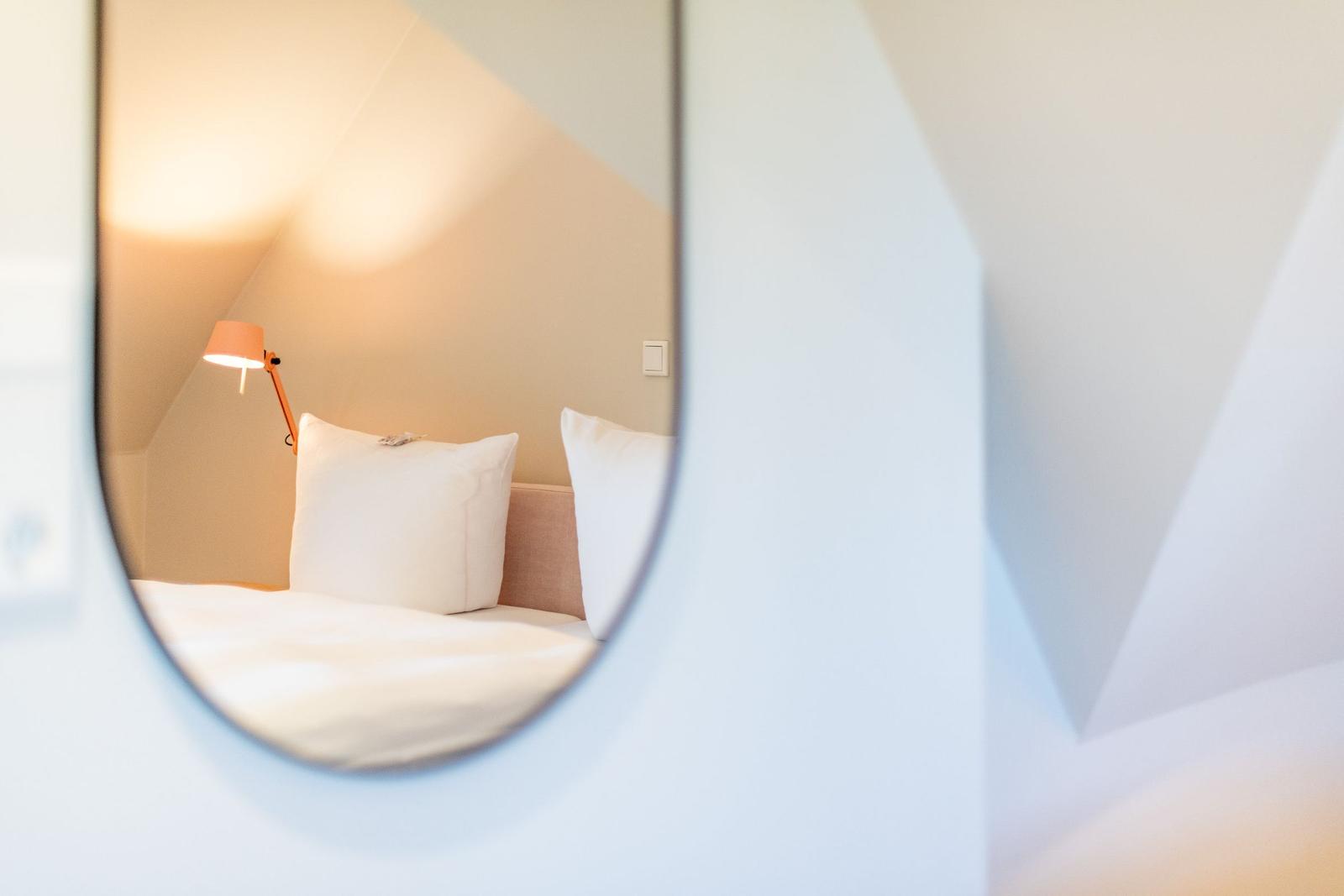 Through a round mirror, a bed with pillows and a lamp is visible.