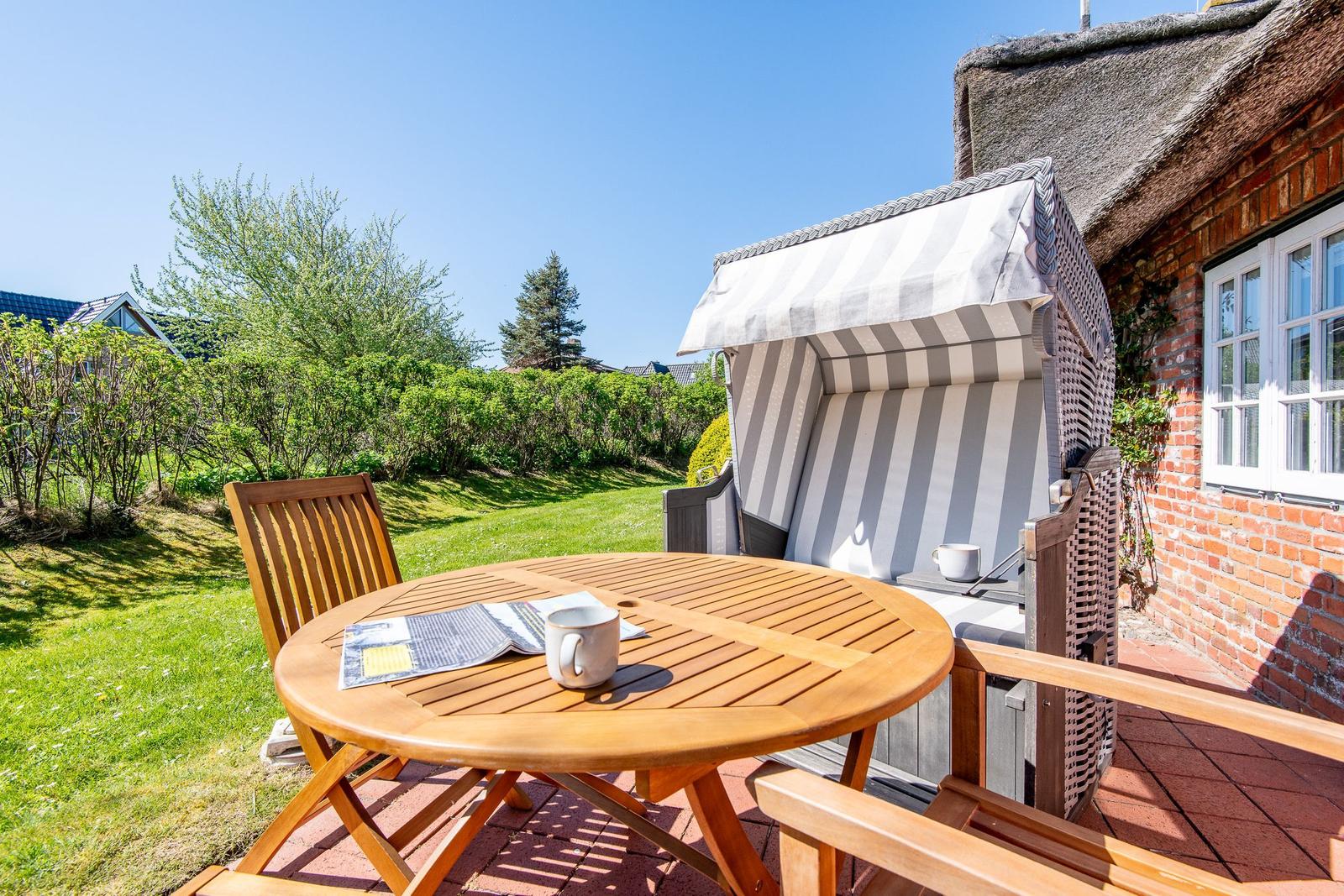 Terrace with wooden table, chair, and beach chair next to a house with thatched roof.