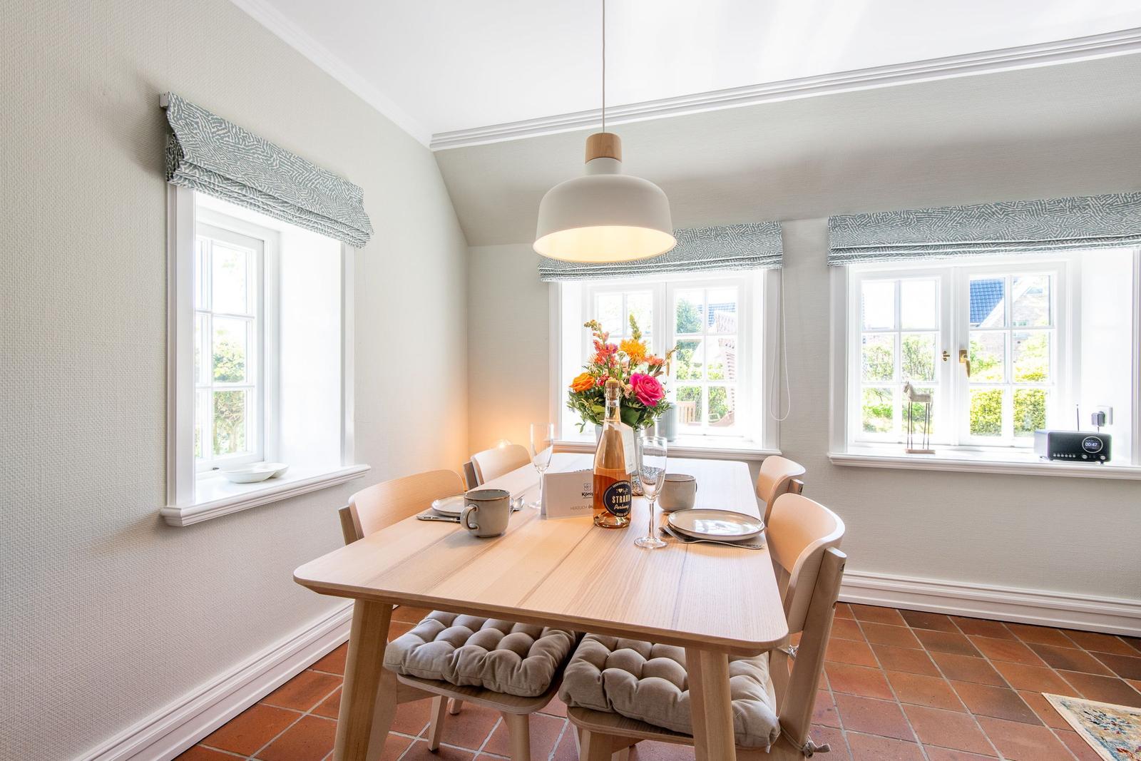 Dining room with wooden table, chairs, and windows. Table set with flowers and bottle.