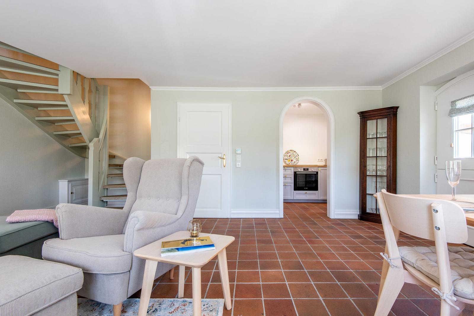 Living area with armchair, table, and terracotta floor. Kitchen and staircase are visible.