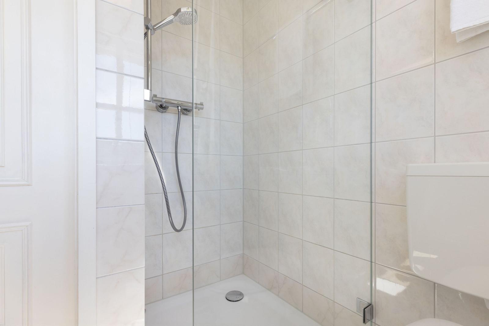 Dusche mit Glaswand und Handbrause, weiße Fliesen und Toilette sichtbar.