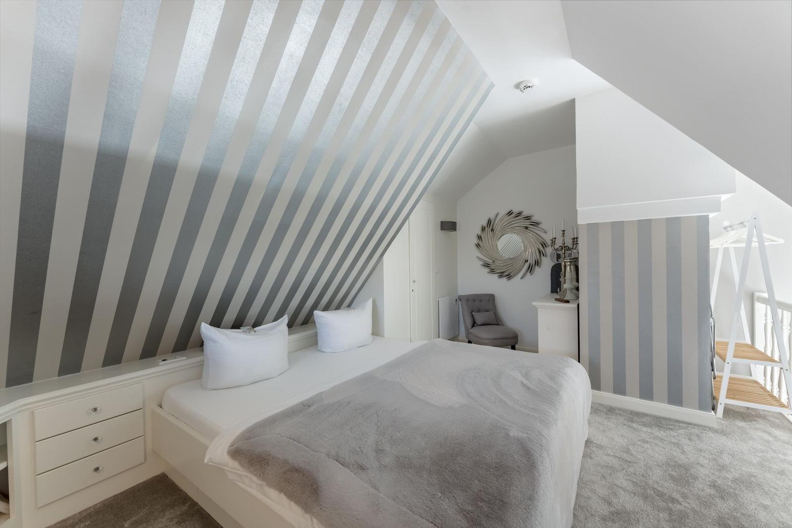 Schlafzimmer mit schräger Decke und gestreifter Wand. Bett, Kleiderschrank und Sessel sind vorhanden.
