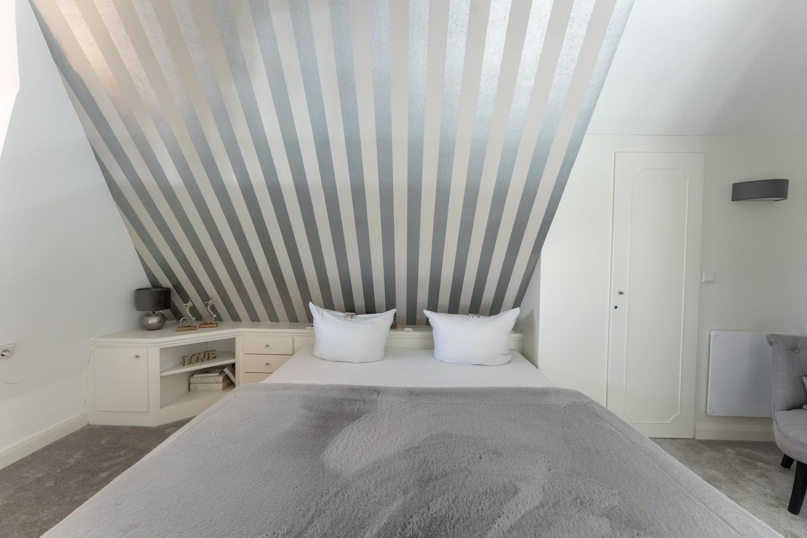 Schlafzimmer mit gestreifter Decke, weißem Bett und grauer Bettdecke.