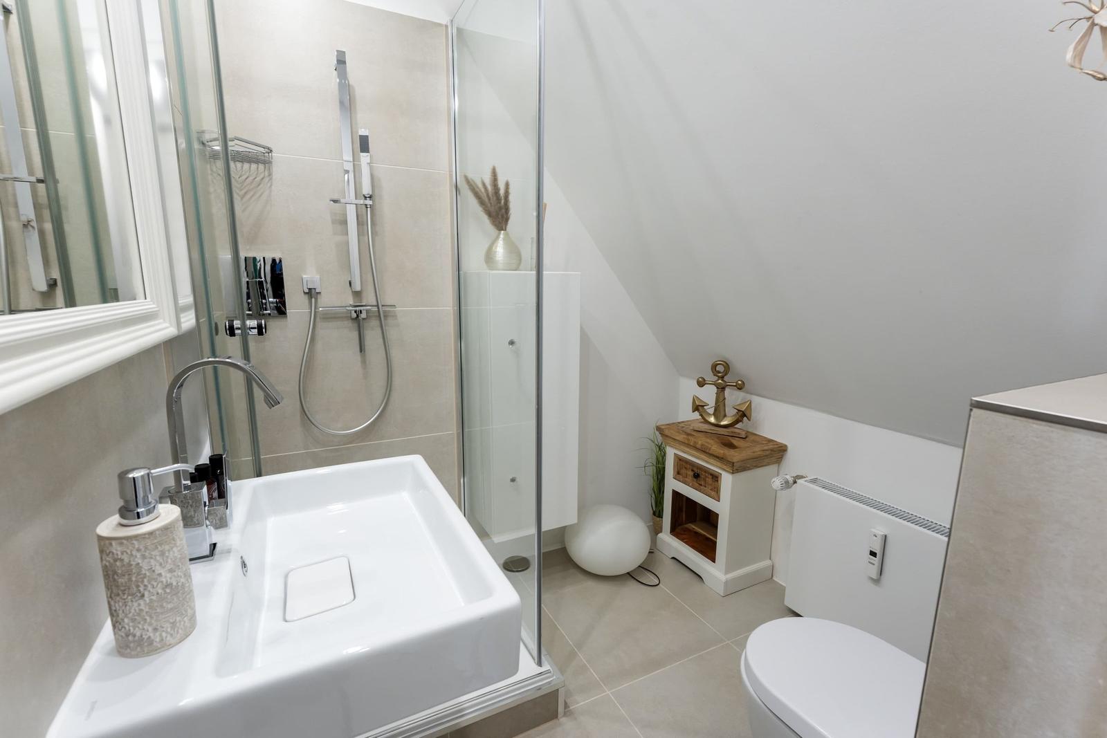 Badezimmer mit Dusche, Waschbecken und Toilette. Holzkommode mit Ankerdekoration.