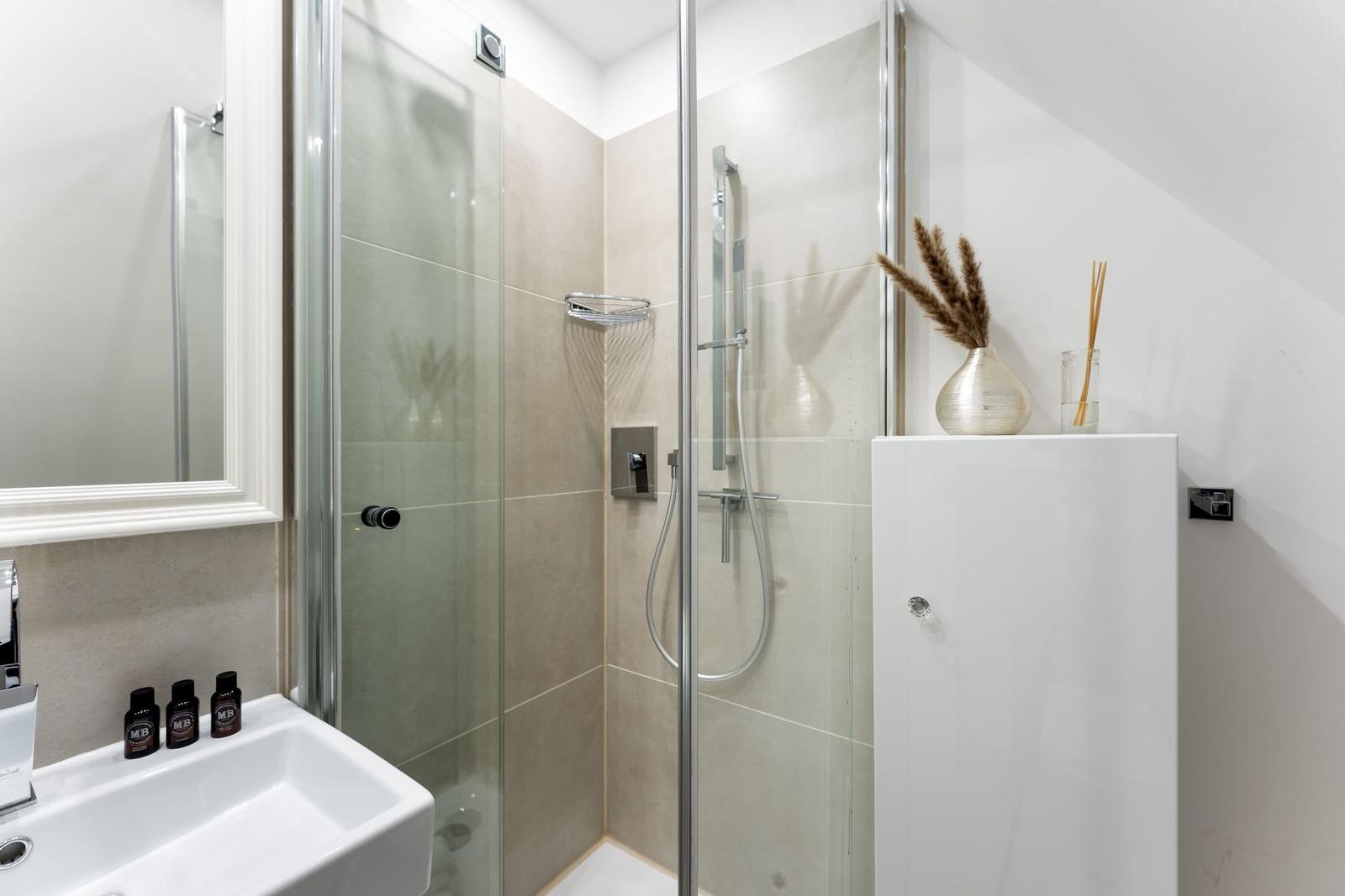 Dusche mit Glaswand, Waschbecken und Spiegel. Dekorative Vase und Duftstäbchen auf Schrank.
