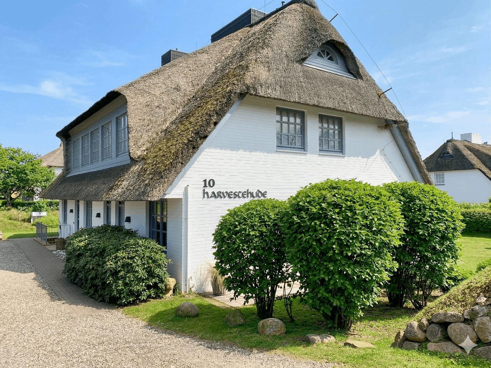 Haus mit strohgedecktem Dach und weißen Wänden, umgeben von grünen Sträuchern.