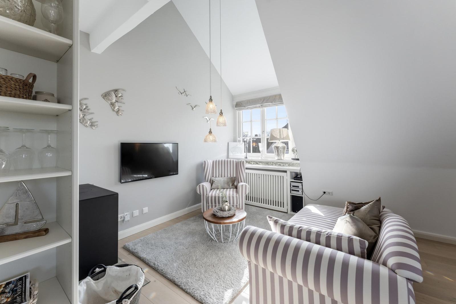 Wohnzimmer mit Sofa, Sessel, Fernseher und Fenster. Regal mit Gläsern und Dekoration.