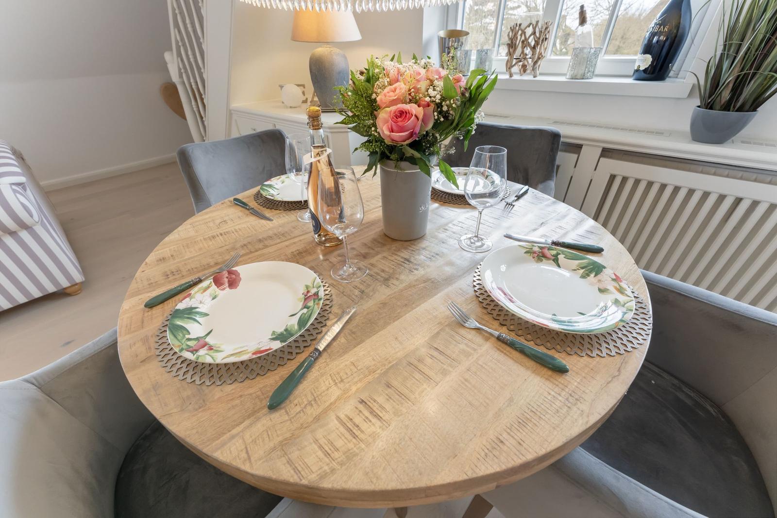 Runder Holztisch mit Platz für vier Personen, Blumenstrauß und Fensterblick.