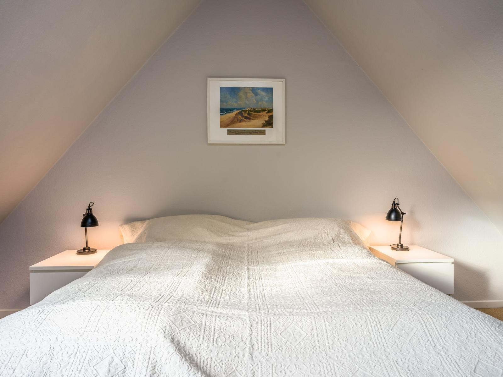Schlafzimmer mit Dachschrägen, Bett mit weißer Bettwäsche und zwei Nachttischen mit Lampen.