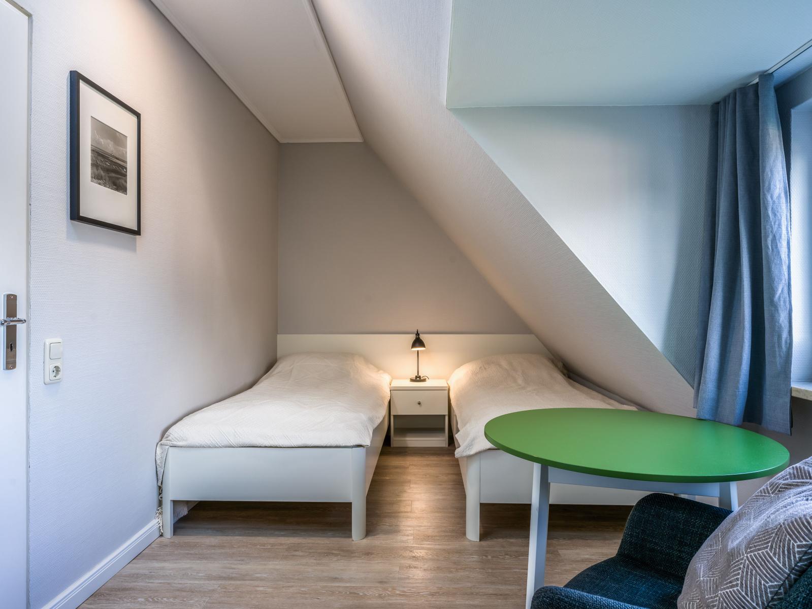 Zwei Einzelbetten, Nachttisch, grüner Tisch und Sessel in einem Dachzimmer.