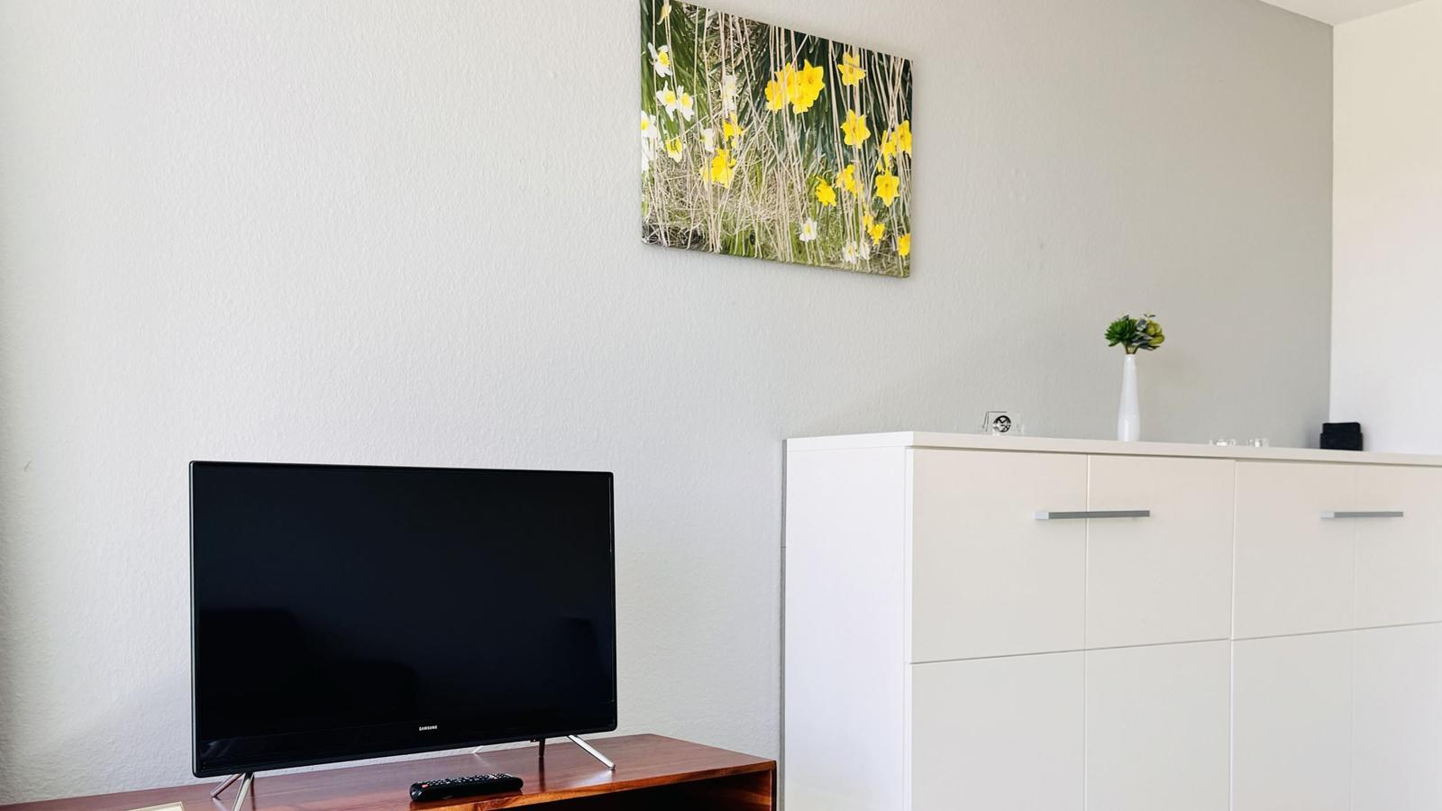 Flachbildfernseher auf Holzregal, weiße Schrankwand mit Vase und Wandbild mit gelben Blumen.