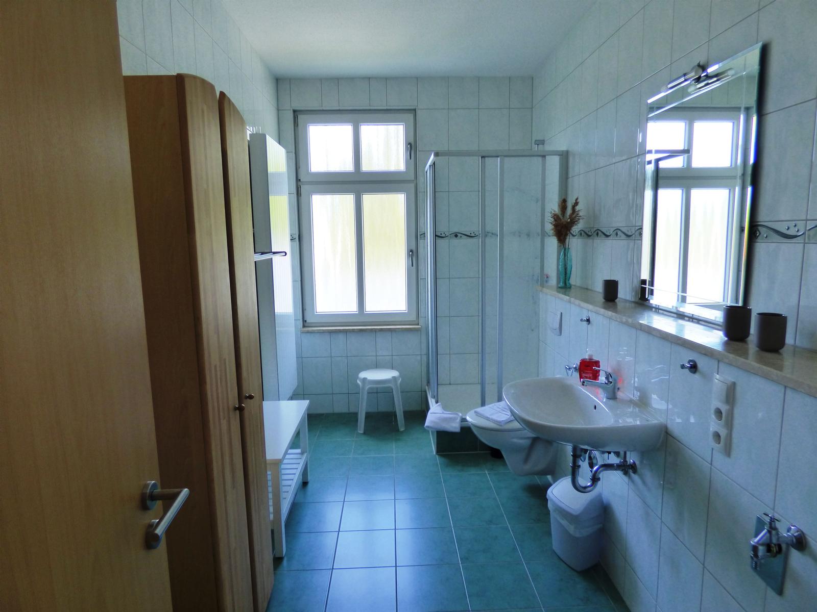 Badezimmer mit Dusche, Waschbecken und Toilette. Fenster und Spiegel sichtbar.