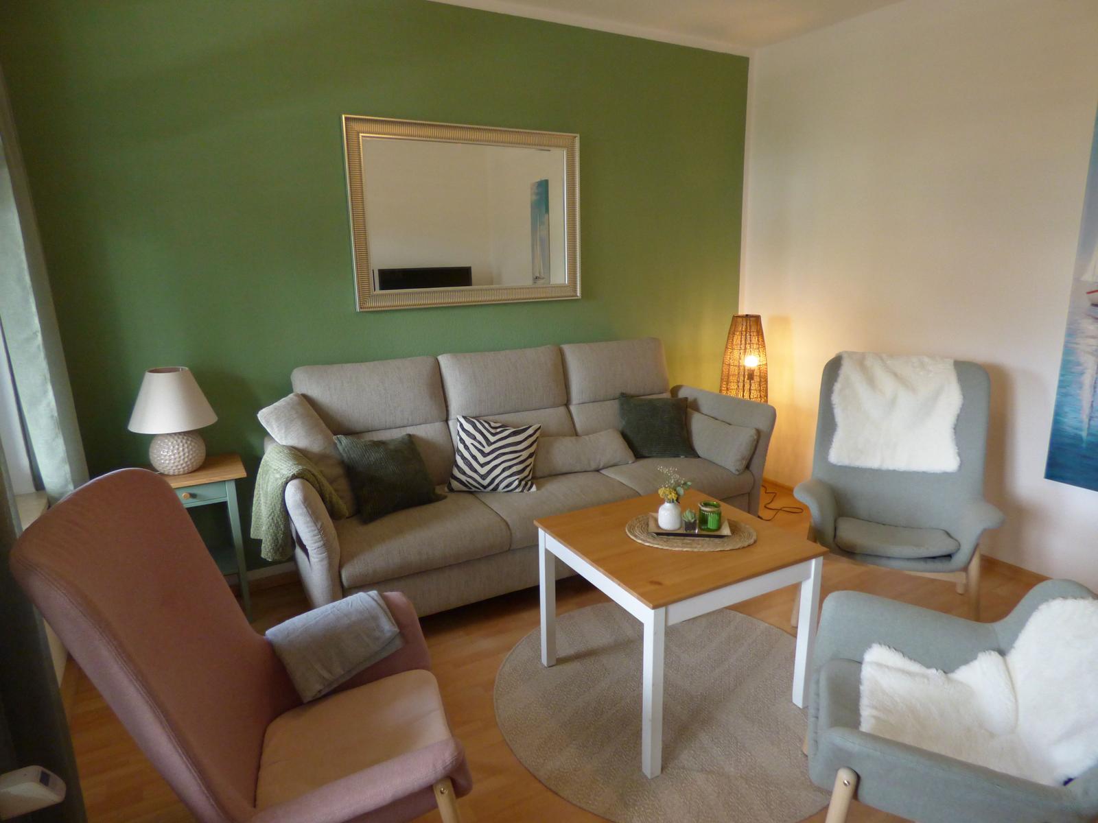 Wohnzimmer mit Sofa, zwei Sesseln und Tisch. Grüne Wand mit Spiegel.