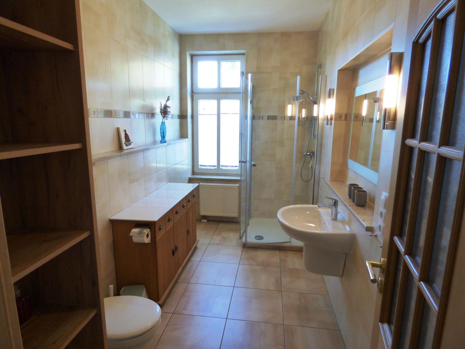 Badezimmer mit Dusche, Waschbecken und Toilette. Holzregal und Fenster sichtbar.