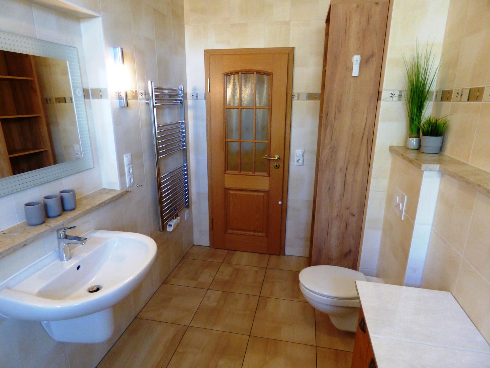 Badezimmer mit Waschbecken, Toilette und Heizung. Holztür und Pflanzen sind sichtbar.