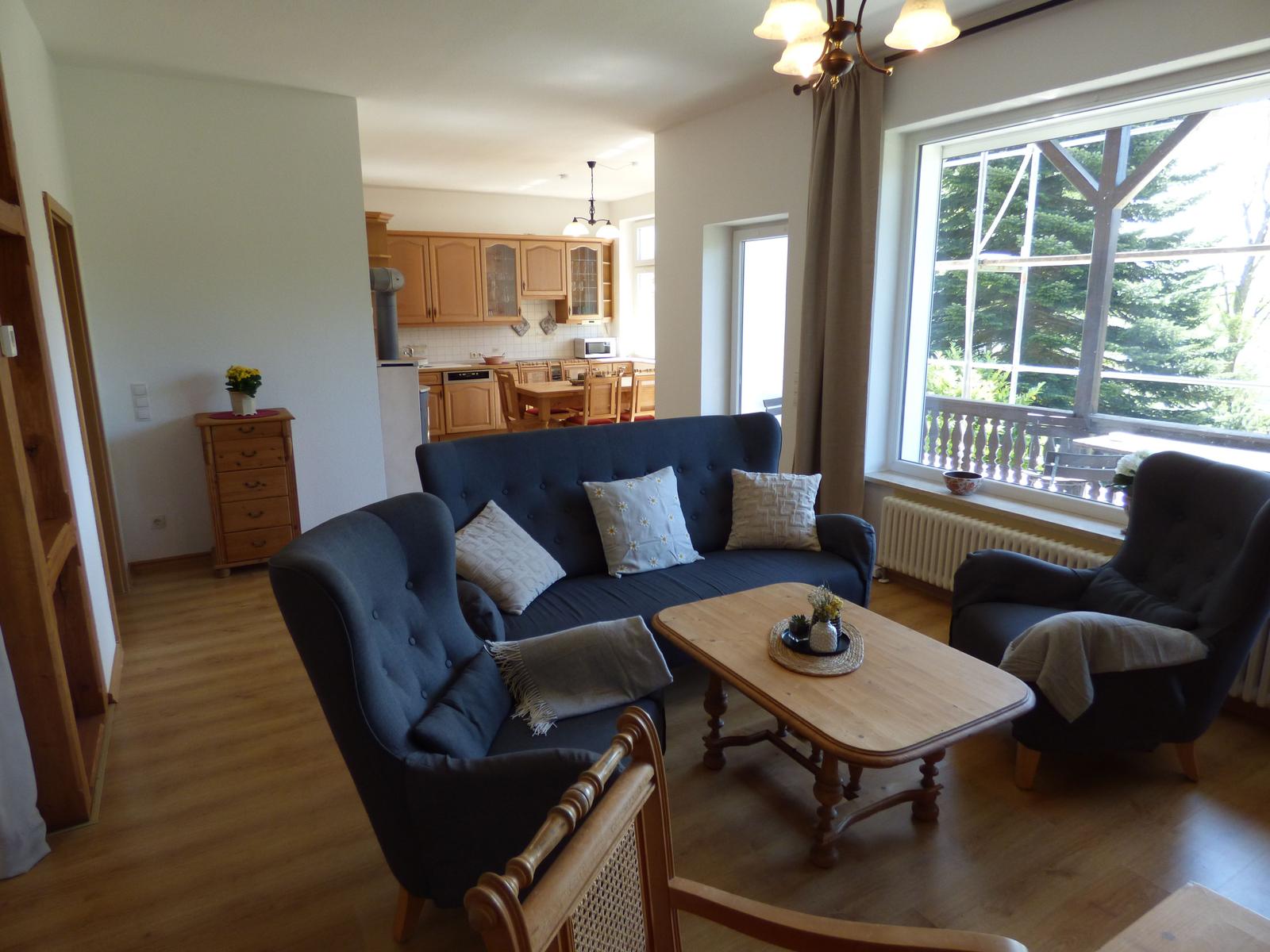 Wohnzimmer mit Sofa, zwei Sesseln und Holztisch. Küche im Hintergrund. Blick auf Balkon mit Bäumen.