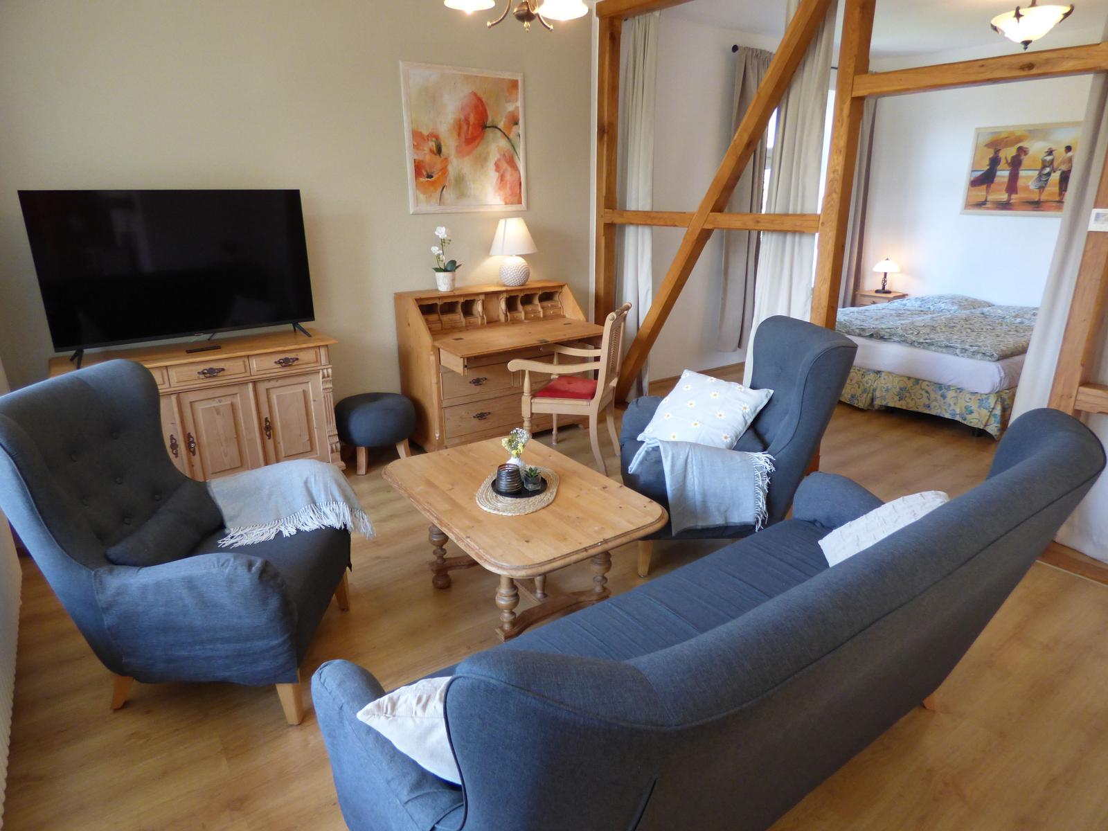 Wohnzimmer mit Sofa, Sessel und Fernseher. Im Hintergrund ist ein Schlafzimmer sichtbar.