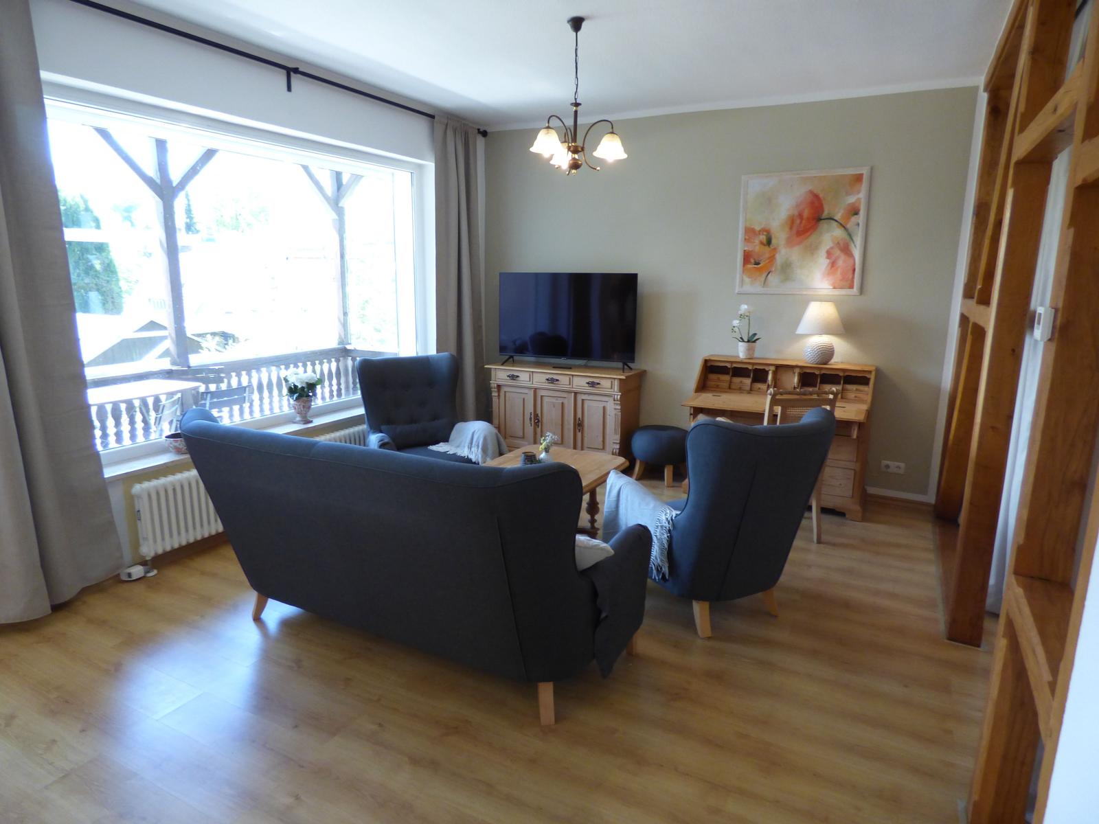Wohnzimmer mit Sofa, Sesseln, Fernseher und Holzmöbeln. Große Fenster mit Blick auf Balkon.