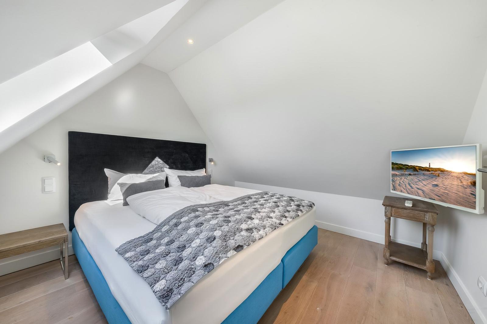 Schlafzimmer mit Doppelbett, TV und Holzboden unter schrägen Dachdecken.