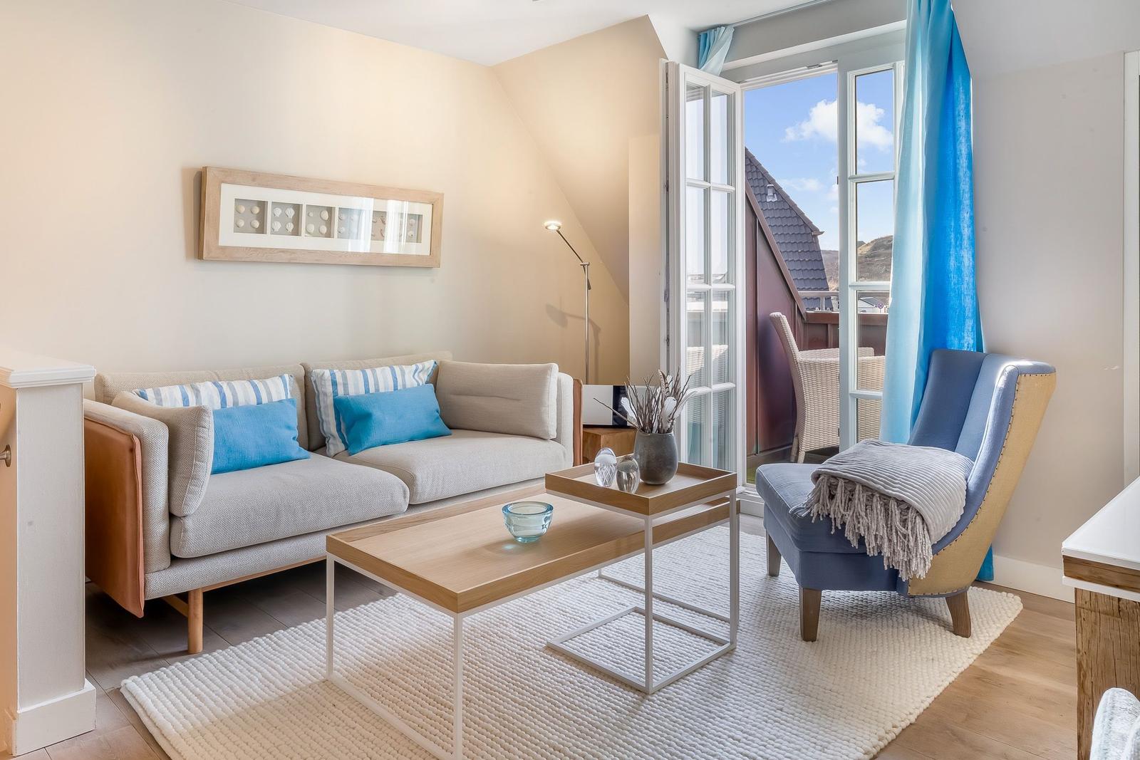 Wohnzimmer mit Sofa, Sessel und Tisch. Durchgehende Glastür führt auf Balkon.