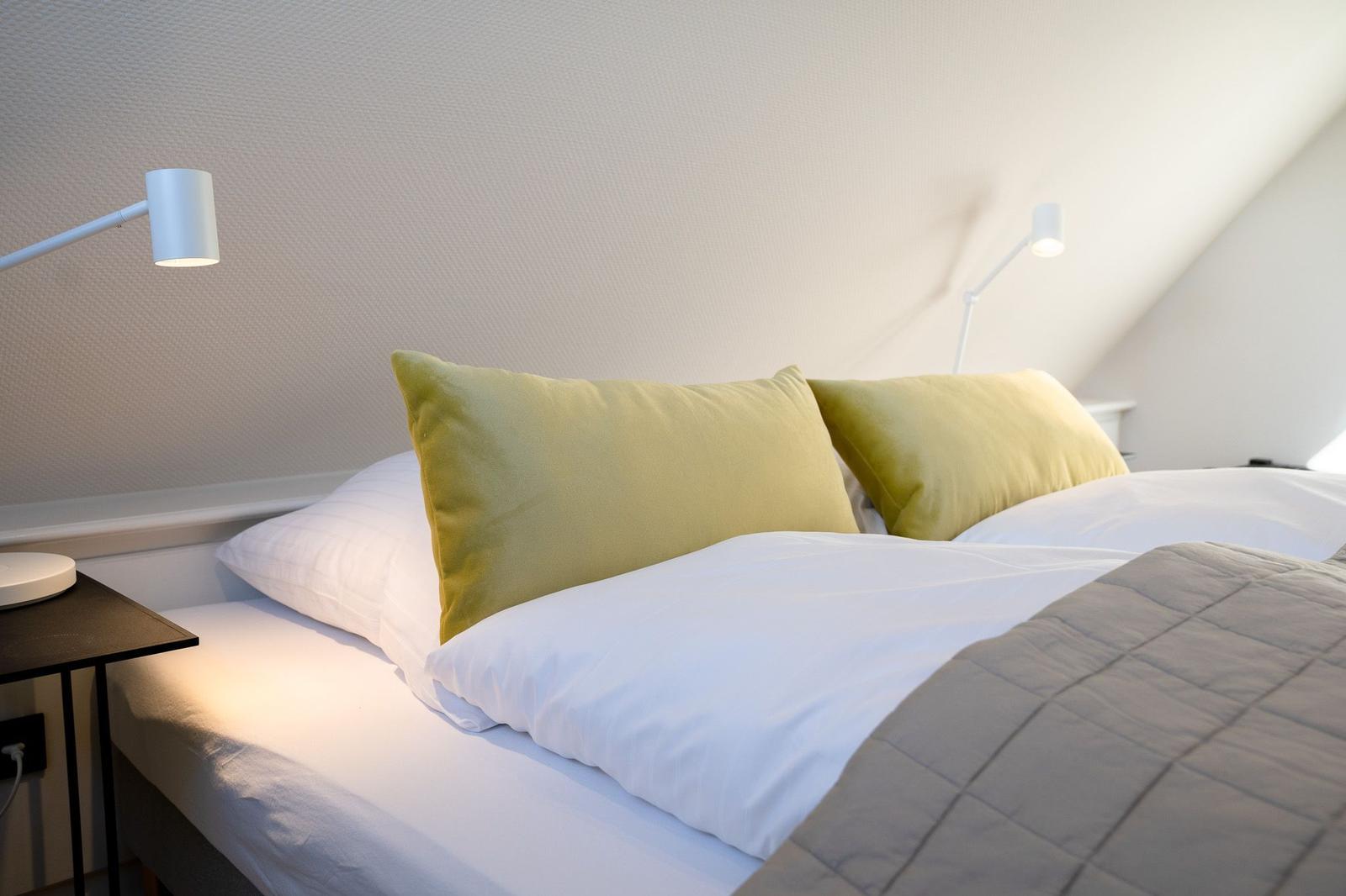 Ein Bett mit weißen Laken und zwei grünen Kissen. An der Wand hängen zwei weiße Stehlampen.