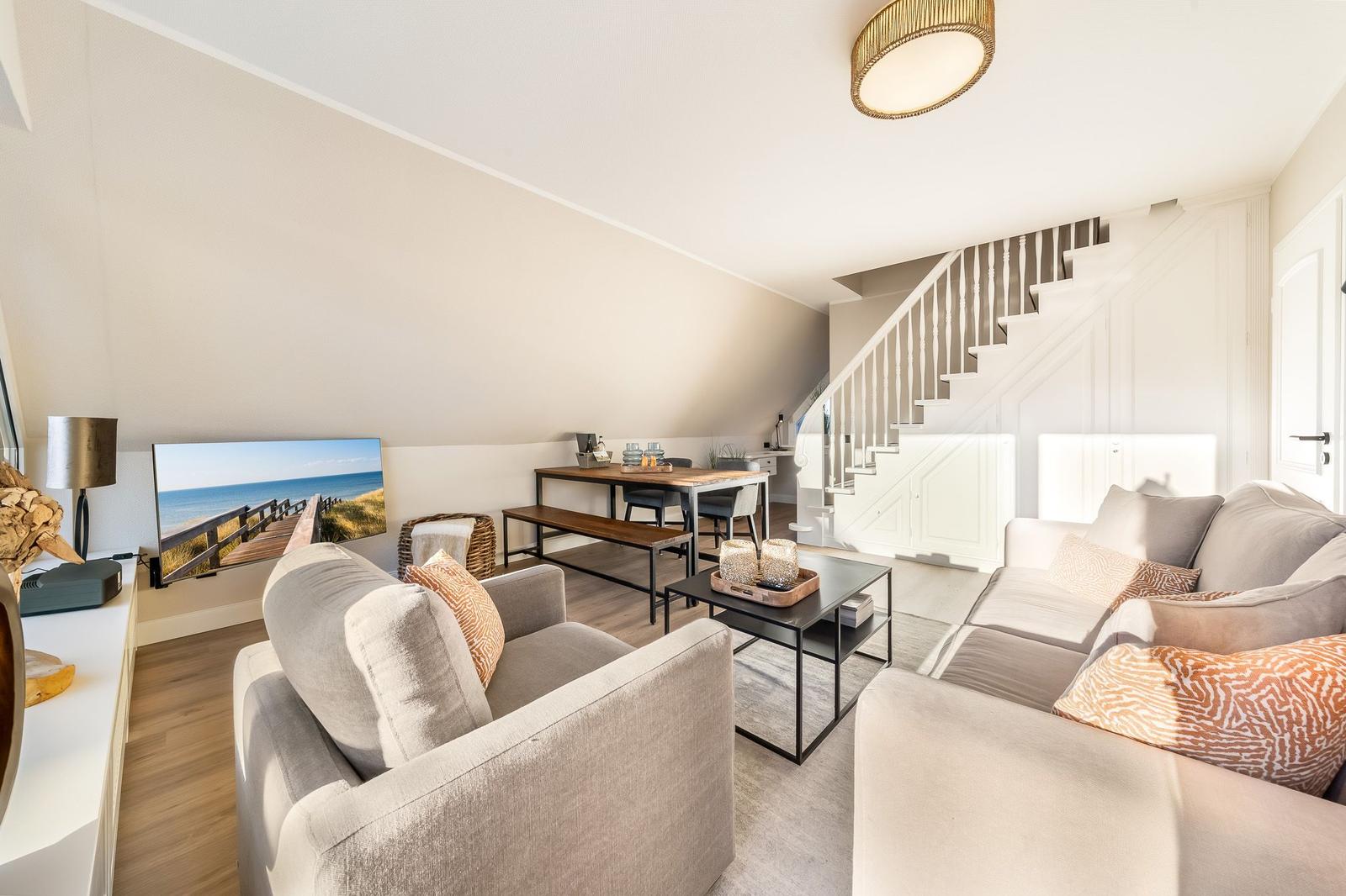 Wohnbereich mit Sofa, Sessel und Essbereich unter Dachschräge. Treppenhaus führt nach oben.