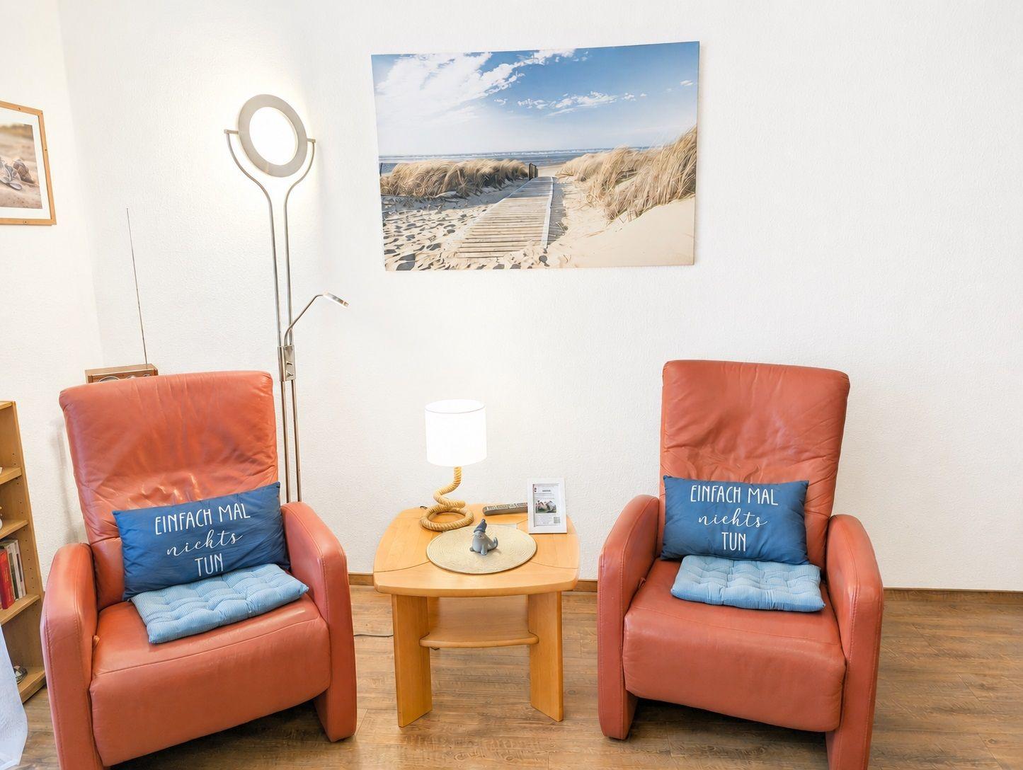 Zwei orangefarbene Sessel mit blauen Kissen stehen um einen Holztisch herum. An der Wand hängt ein Strandbild.