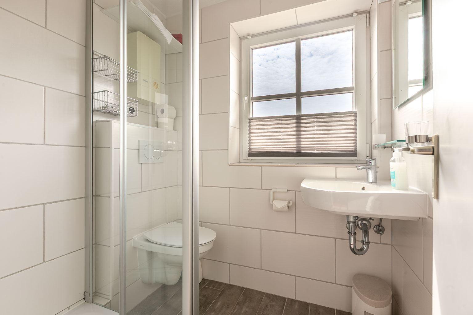 Badezimmer mit Dusche, Toilette, Waschbecken und Fenster mit Jalousie.