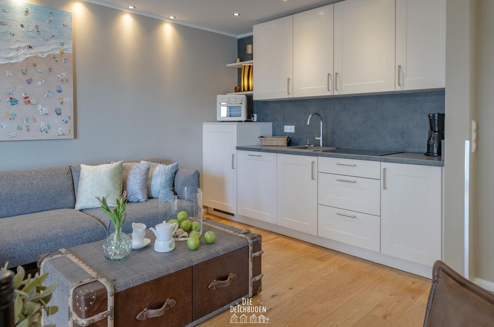 Wohnbereich mit Sofa, Kühlschrank und Küchenzeile. Holzboden und Wandbild mit Strandmotiv.
