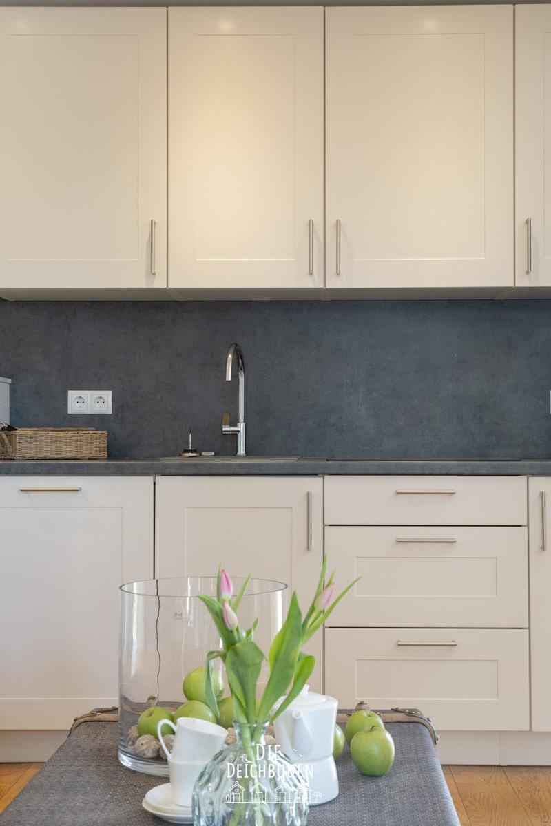 Küche mit weißen Schränken, grauer Rückwand und Arbeitsplatte. Tisch mit Blumen und Äpfeln.
