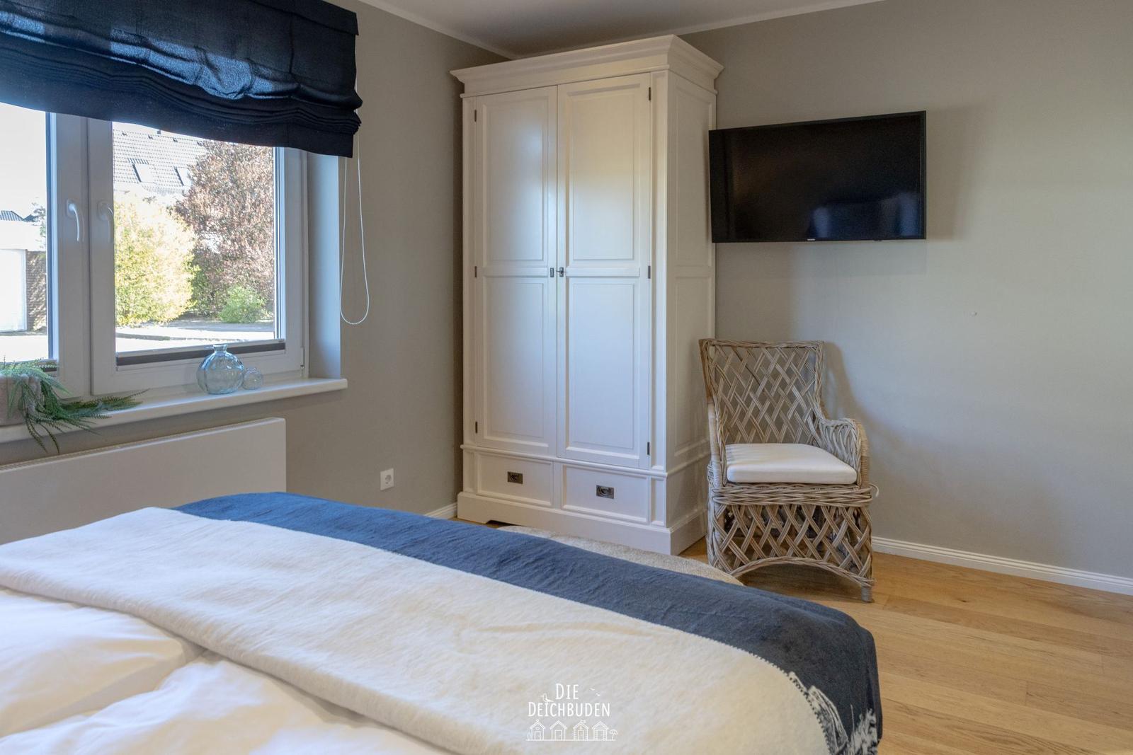Schlafzimmer mit Bett, Kleiderschrank, Sessel und Wandfernseher. Fenster mit Blick nach draußen.
