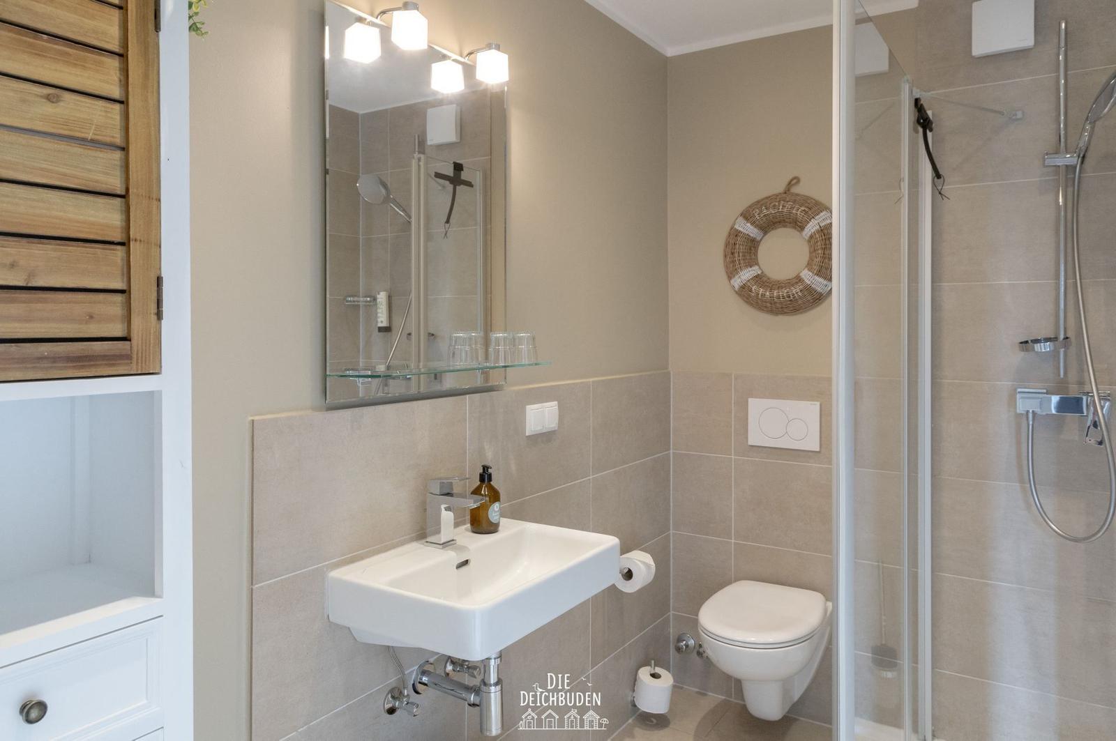 Badezimmer mit Dusche, Waschbecken und Toilette. Holzschrank und Spiegel mit Licht.